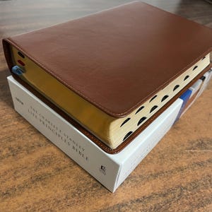 May include: A brown leather-bound Bible with gold-edged pages rests on a white box. The box features the text "THE CHARLES F. STANLEY LIVE PRINCIPLES BIBLE." The Bible has black page markers and a ribbon bookmark.