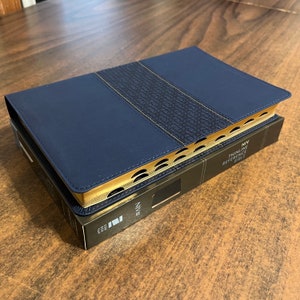 May include: A navy blue leather-bound Bible with gold accents and a decorative pattern on the spine. The cover reads "NIV Thinline Reference Bible".