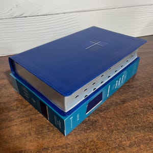 May include: A blue NIV Quest Study Bible in a personal size, resting on its matching blue and teal cardboard box. The Bible cover features a simple cross emblem, and the box displays the text NIV Quest Study Bible Personal Size in white.