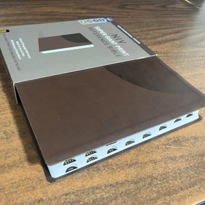 May include: A brown leather-bound NIV Super Giant Print Reference Bible with labeled tabs. The book is partially open, revealing the pages and the text "NIV Super Giant Print Reference Bible" on the cover.