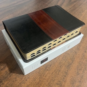 May include: A black and brown leather-bound book with gold-edged pages rests on a white box. The book has a brown stripe down the center and is likely a Bible or similar religious text. The book is closed and appears new.