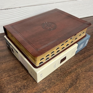 May include: A brown leather-bound King James Version (KJV) Bible with gold-gilded edges and a Celtic knot design on the cover. The Bible is closed and sitting on a white box with the words "King James Version" printed on it.