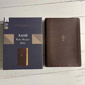 May include: A brown leather-bound Bible with a gold cross embossed on the cover. The Bible is in a blue box with the text "NASB Wide-Margin Bible" printed on the front.