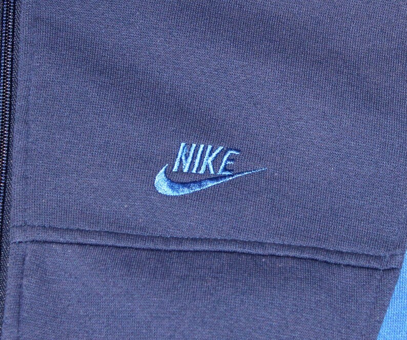 May include: Close-up of a navy blue Nike jacket. The Nike logo is embroidered in light blue on the upper left chest. The jacket has a full zipper and a contrasting light blue panel on the right side.