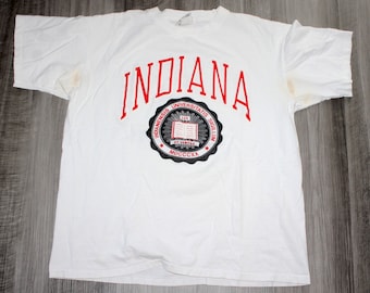 Könnte beinhalten: Weißes T-Shirt mit einem roten "INDIANA"-Aufdruck und einem schwarzen und weißen Siegel mit dem Text "INDIANAENSIS UNIVERSITATIS SIGILLUM LUX ET VERITAS MDCCCX",