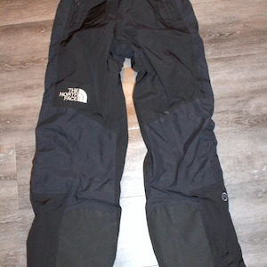 Puede incluir: Pantalones de nieve negros con el logo blanco "THE NORTH FACE" en la pierna izquierda. Los pantalones tienen paneles reforzados en las rodillas y la parte inferior de las piernas. Los pantalones tienen una etiqueta "GORE-TEX".