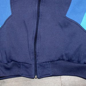 May include: Close-up of a vintage zip-up sweatshirt in shades of blue. The jacket features a navy blue body with light and medium blue color block accents on the sleeves. The bottom hem and cuffs are ribbed.