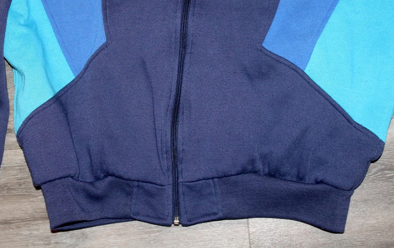 May include: Close-up of a vintage zip-up sweatshirt in shades of blue. The jacket features a navy blue body with light and medium blue color block accents on the sleeves. The bottom hem and cuffs are ribbed.