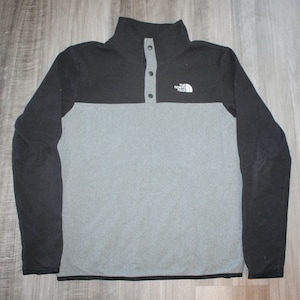 May include: A gray and black long-sleeve fleece pullover with a snap button closure at the neck. The North Face logo is on the upper right side of the shirt.