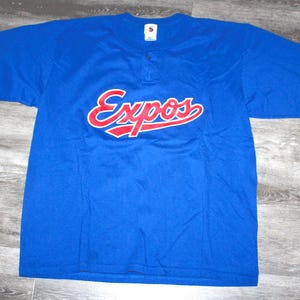 Può includere: Una maglia da baseball blu degli Expos con la scritta "Expos" in rosso con un contorno bianco sul petto. La maglia ha uno scollo con bottoni ed è realizzata in un tessuto traspirante.