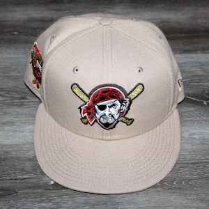 Pode incluir: Boné de beisebol bege com aba reta. A frente apresenta um logotipo da equipe de um pirata com uma bandana vermelha e bastões cruzados. Um patch colorido está na lateral. O boné tem um ajuste estruturado.