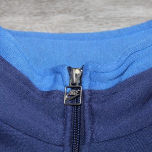 May include: Close-up of a blue Nike track jacket collar. The collar is two-toned, with a lighter blue interior and a darker blue exterior. A silver zipper with the Nike logo is visible.