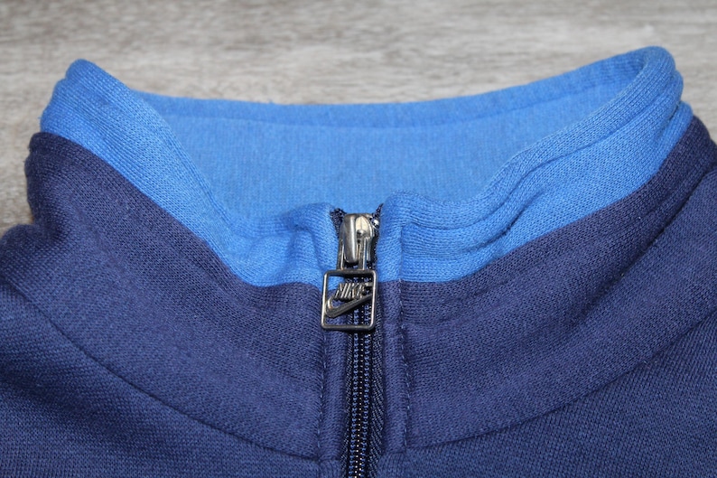 May include: Close-up of a blue Nike track jacket collar. The collar is two-toned, with a lighter blue interior and a darker blue exterior. A silver zipper with the Nike logo is visible.