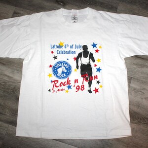 May include: White t-shirt with a blue, red and yellow graphic design. The graphic features a runner, the text "Latrobe 4th of July Celebration", "Rolling Rock", "Rock 'n Run", and "5 Mile '98".