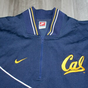 Pu&ograve; includere: Una giacca blu navy con zip e bordini bianchi e dorati. La giacca ha un logo Nike sul colletto e un logo "Cal" ricamato in oro sul davanti.