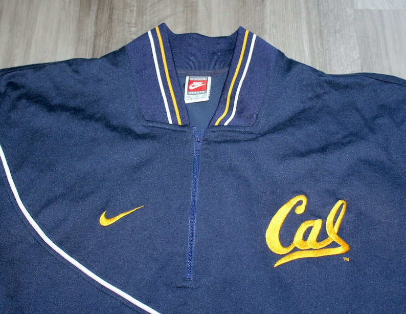 Pu&ograve; includere: Una giacca blu navy con zip e bordini bianchi e dorati. La giacca ha un logo Nike sul colletto e un logo "Cal" ricamato in oro sul davanti.