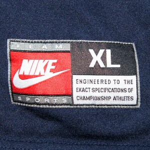 Pu&ograve; includere: Un'etichetta Nike Team Sports blu navy con cuciture bianche. L'etichetta presenta il logo Nike swoosh in bianco su sfondo rosso. I testi "TEAM" e "SPORTS" sono in bianco su sfondo grigio. Il testo "NIKE" &egrave; in bianco su sfondo rosso. Il testo "XL" &egrave; in bianco su sfondo nero. Il testo "ENGINEERED TO THE EXACT SPECIFICATIONS OF CHAMPIONSHIP ATHLETES" &egrave; in bianco su sfondo grigio.