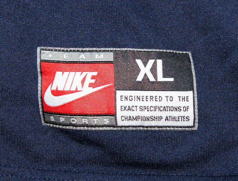 Pu&ograve; includere: Un'etichetta Nike Team Sports blu navy con cuciture bianche. L'etichetta presenta il logo Nike swoosh in bianco su sfondo rosso. I testi "TEAM" e "SPORTS" sono in bianco su sfondo grigio. Il testo "NIKE" &egrave; in bianco su sfondo rosso. Il testo "XL" &egrave; in bianco su sfondo nero. Il testo "ENGINEERED TO THE EXACT SPECIFICATIONS OF CHAMPIONSHIP ATHLETES" &egrave; in bianco su sfondo grigio.