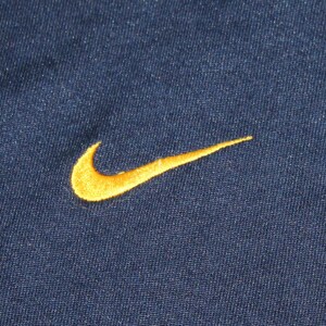 Pu&ograve; includere: Un tessuto blu navy con un logo Nike swoosh ricamato in oro.