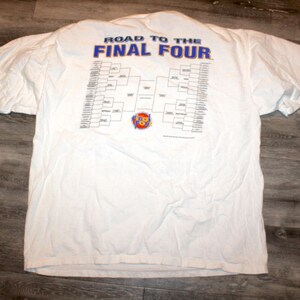 Pu&ograve; includere: T-shirt bianca con la scritta blu "ROAD TO THE FINAL FOUR" e un tabellone. La maglietta ha maniche corte ed &egrave; appoggiata su una superficie di legno.