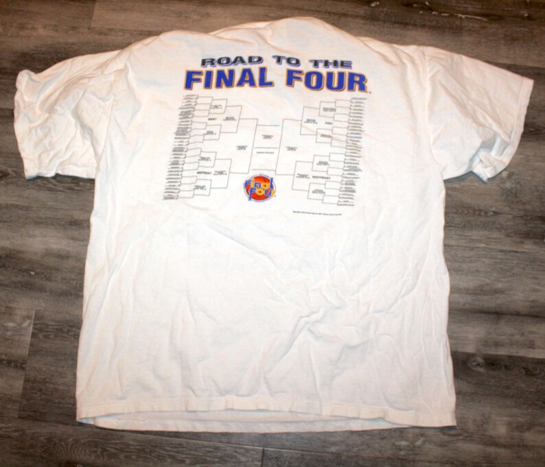 Pu&ograve; includere: T-shirt bianca con la scritta blu "ROAD TO THE FINAL FOUR" e un tabellone. La maglietta ha maniche corte ed &egrave; appoggiata su una superficie di legno.