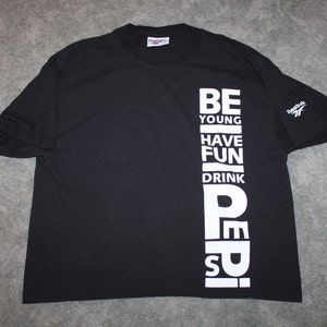 Op de afbeelding: Zwart T-shirt met witte tekst die "Be Young Have Fun Drink Pepsi" op de voorkant staat. Het shirt heeft een Reebok-logo op de rechtermouw.
