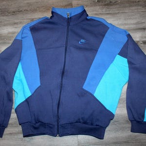 May include: A vintage Nike track jacket in shades of blue. The jacket features a full zip, a stand-up collar, and the Nike logo embroidered on the chest. The color-blocked design includes navy, medium blue, and turquoise panels.
