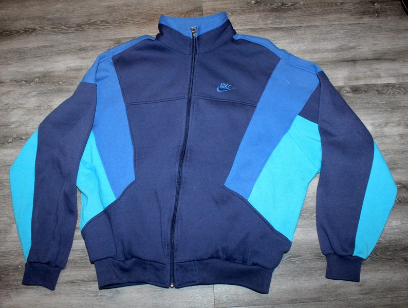 May include: A vintage Nike track jacket in shades of blue. The jacket features a full zip, a stand-up collar, and the Nike logo embroidered on the chest. The color-blocked design includes navy, medium blue, and turquoise panels.