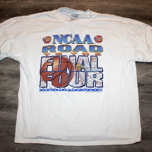 Pu&ograve; includere: T-shirt bianca con la scritta "NCAA Road to the Final Four Indianapolis" in blu, arancione e marrone. Il design include una grafica di basket e una mappa.