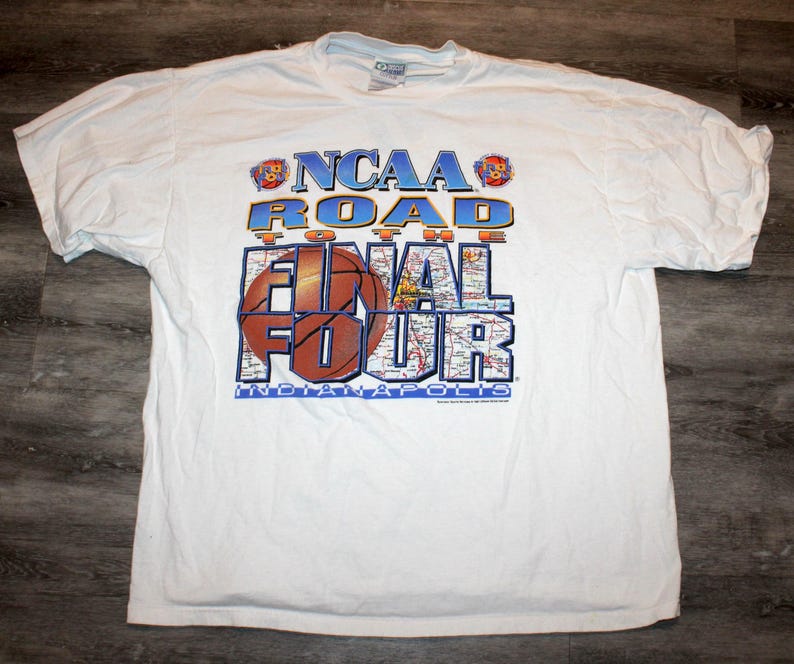Pu&ograve; includere: T-shirt bianca con la scritta "NCAA Road to the Final Four Indianapolis" in blu, arancione e marrone. Il design include una grafica di basket e una mappa.