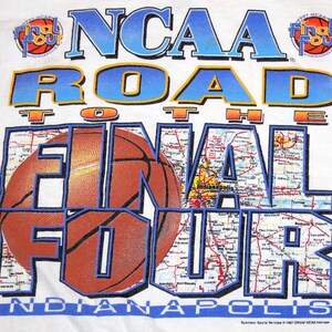 Pu&ograve; includere: T-shirt bianco con un design a tema basket. Il testo recita "NCAA Road to the Final Four Indianapolis". Il design incorpora una grafica di pallone da basket e uno sfondo di mappa, con i loghi "1997" e "Final Four".