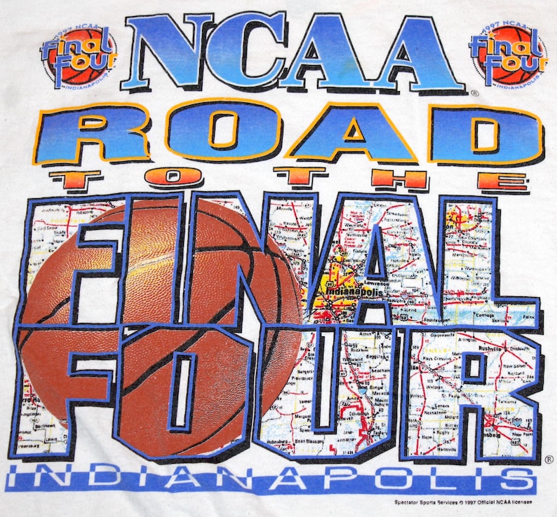 Pu&ograve; includere: T-shirt bianco con un design a tema basket. Il testo recita "NCAA Road to the Final Four Indianapolis". Il design incorpora una grafica di pallone da basket e uno sfondo di mappa, con i loghi "1997" e "Final Four".