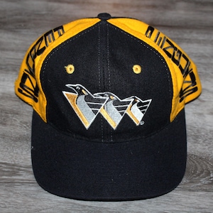 Puede incluir: Gorra de béisbol negra y amarilla con la palabra "PENGUINS" en el borde. La gorra presenta un logotipo de tres pingüinos dentro de un triángulo blanco y amarillo. La gorra está hecha de una tela duradera.