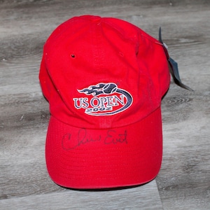 Op de afbeelding: Een rode baseballpet met het US Open 2002 logo geborduurd op de voorkant. De pet is gesigneerd met "Chris Evert" in zwarte stift.