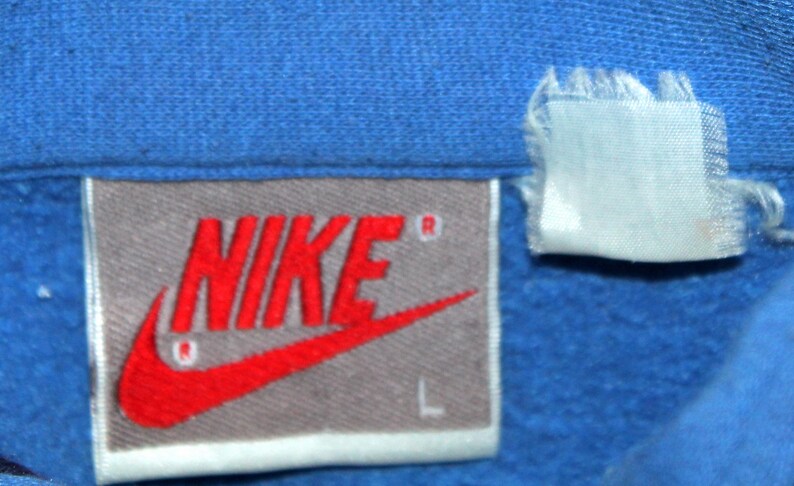 May include: Close-up of a blue garment with a Nike label. The label is rectangular with the Nike logo in red and the brand name in red. The size "L" is printed in white. A white tag is attached to the garment.