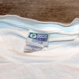Pu&ograve; includere: Primo piano di una maglietta bianca di cotone con scollo tondo. La maglietta ha un'etichetta di Discus Athletic, con un logo verde e blu e la scritta "COTTON". L'etichetta indica anche la taglia 2XL.