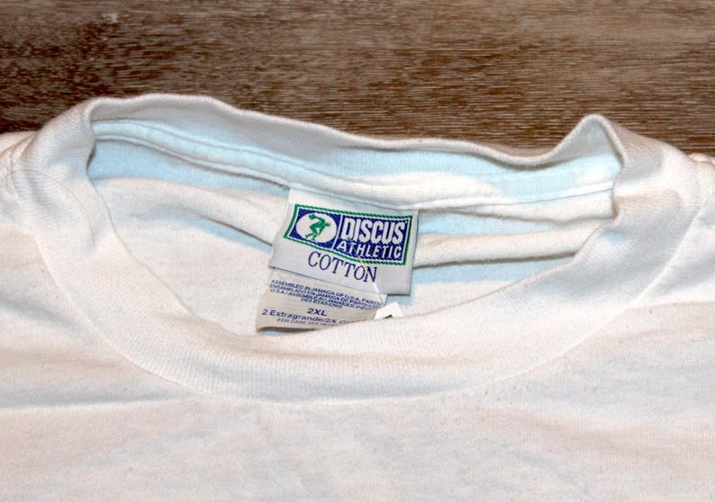 Pu&ograve; includere: Primo piano di una maglietta bianca di cotone con scollo tondo. La maglietta ha un'etichetta di Discus Athletic, con un logo verde e blu e la scritta "COTTON". L'etichetta indica anche la taglia 2XL.
