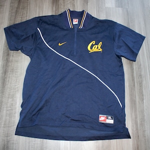 Pu&ograve; includere: Una polo a maniche corte blu navy con zip bianca e bordini bianchi. La polo ha un logo "Cal" dorato sul davanti e un logo Nike sul petto sinistro. La polo &egrave; taglia XL.