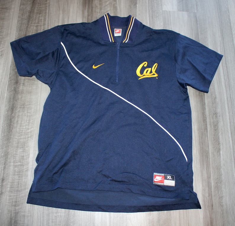 Pu&ograve; includere: Una polo a maniche corte blu navy con zip bianca e bordini bianchi. La polo ha un logo "Cal" dorato sul davanti e un logo Nike sul petto sinistro. La polo &egrave; taglia XL.