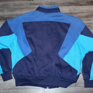 May include: A vintage track jacket in shades of blue. The jacket features a full zipper, a stand-up collar, and color-blocked panels in navy, medium blue, and turquoise. The cuffs and waistband are ribbed.