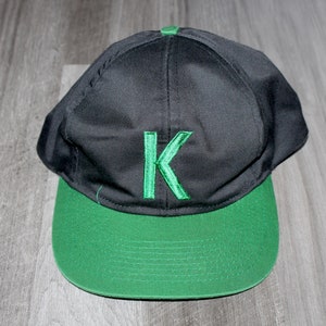 Könnte beinhalten: Eine schwarze und grüne Baseballkappe mit einem grünen, gestickten Buchstaben "K" auf der Vorderseite.