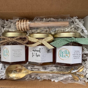 May include: A gift box containing three jars of honey with decorative labels and ribbons, a wooden honey dipper, and a gold-colored spoon. The jars have the text "Peace begins with tea", "meant to bee", and "Bella Mae's Tea Co."