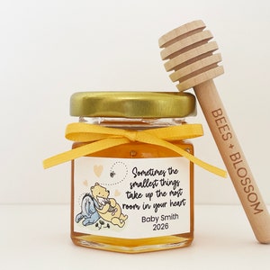 May include: A small glass jar filled with honey, tied with a yellow ribbon, and a wooden honey dipper with the text "BEES + BLOSSOM" on it. The jar has a label with a Winnie the Pooh illustration and the text "Sometimes the smallest things take up the most room in your heart. Baby Smith 2026."
