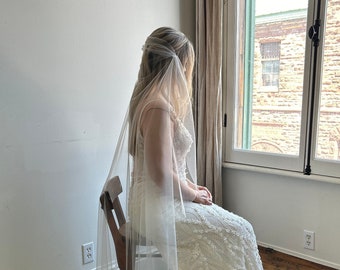 May include: A floor-length ivory wedding dress with intricate white floral lace embroidery and a long train, paired with a sheer, translucent tulle veil that drapes over the shoulders and extends down the back, creating a layered, elegant silhouette.