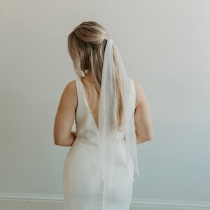 May include: A bride wearing a white wedding dress with a long train and a white veil. The dress has a low back and a button-up closure.