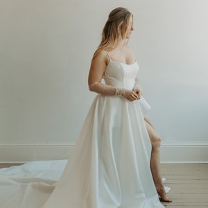 Pu&ograve; includere: Abito da sposa bianco con gonna fluida e spacco alto. L'abito presenta un corpetto aderente con spalline sottili. La sposa indossa guanti trasparenti con dettagli di perle. L'abito ha un lungo strascico.