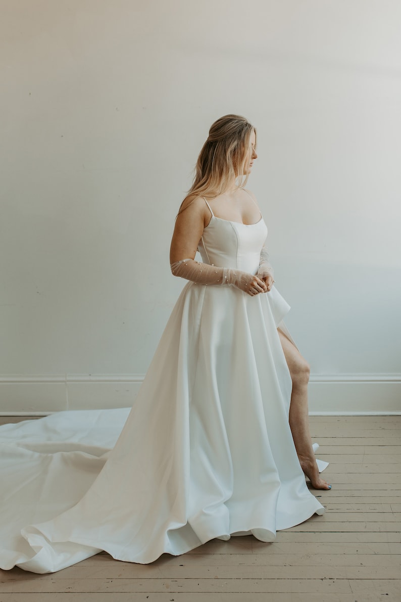 Pu&ograve; includere: Abito da sposa bianco con gonna fluida e spacco alto. L'abito presenta un corpetto aderente con spalline sottili. La sposa indossa guanti trasparenti con dettagli di perle. L'abito ha un lungo strascico.