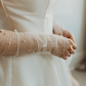 Pu&ograve; includere: Primo piano di un abito da sposa bianco con guanti lunghi e trasparenti. I guanti sono adornati con perle sparse. L'abito ha un design semplice ed elegante. L'estetica generale &egrave; morbida e romantica, adatta all'abbigliamento da sposa.