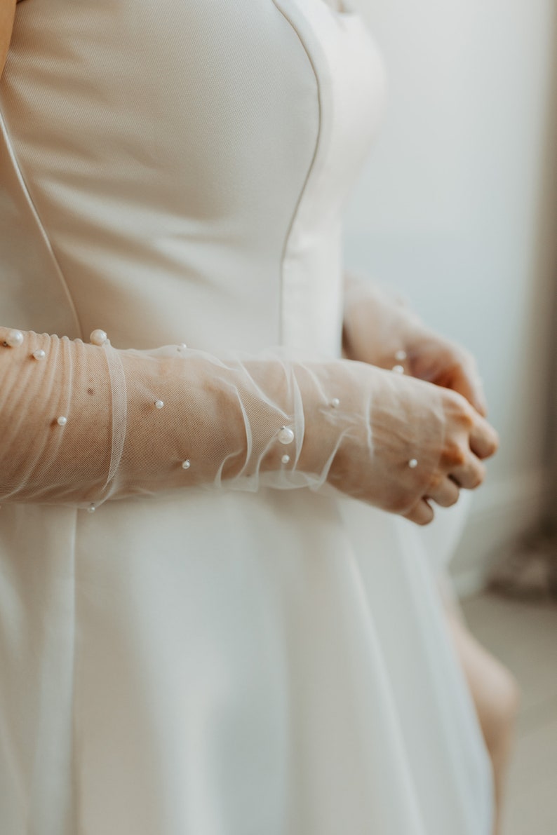 Pu&ograve; includere: Primo piano di un abito da sposa bianco con guanti lunghi e trasparenti. I guanti sono adornati con perle sparse. L'abito ha un design semplice ed elegante. L'estetica generale &egrave; morbida e romantica, adatta all'abbigliamento da sposa.