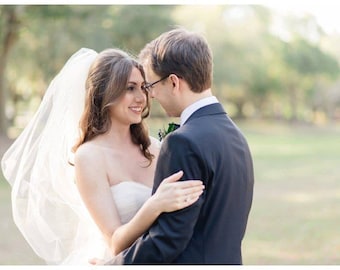 May include: A bride and groom are looking at each other with smiles on their faces. The bride is wearing a white wedding dress and a veil. The groom is wearing a dark suit and a tie. They are standing in a grassy field with trees in the background.